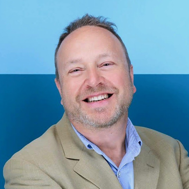 A smiling, fair-skinned man with short brown hair and neatly trimmed beard. He wears a tan blazer over a light blue button-up shirt with an open collar.
